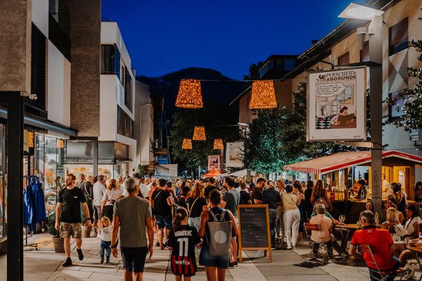 Menschen genießen einen sommerlichen Abend bei den Lana23°° Shoppingnächten in der Fußgängerzone von Lana mit Musik, Essen und Lichtern.