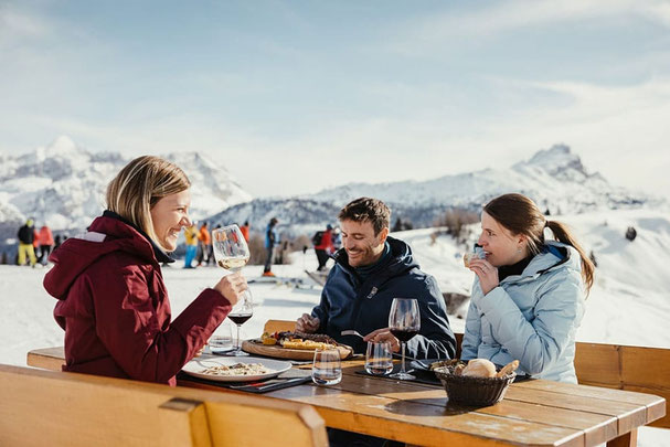 Tre persone si godono pasta e vino su un tavolo di legno in un rifugio innevato in Alta Badia durante il  skisafari, circondati da un panorama invernale soleggiato e dallo scenario delle Dolomiti.