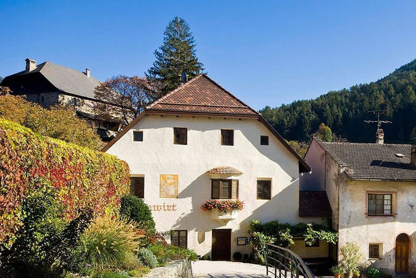 Ristorante Turmwirt a Gudon, storica locanda con facciata bianca, balcone fiorito e ponte, circondata da vigneti e paesaggi autunnali.