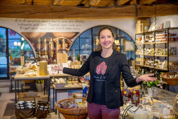 Stefanie, la proprietaria della casa del vino e del gusto Vis à Vis a S. Paolo, presenta una bottiglia di vino tra specialità gastronomiche e prelibatezze in un'accogliente enoteca.