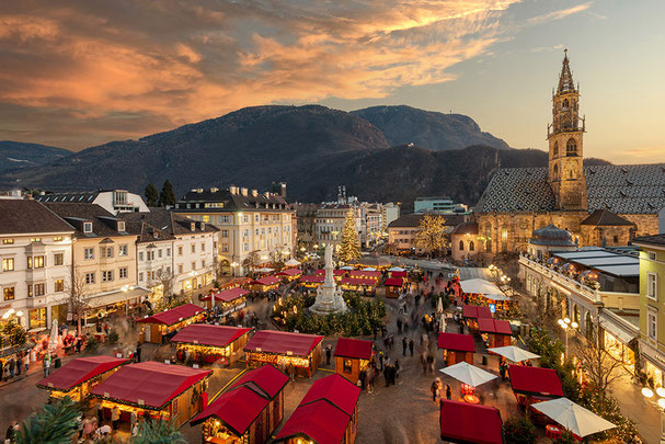 Stimmungsvoller Bozner Christkindlmarkt bei Sonnenuntergang auf dem Waltherplatz, mit festlich beleuchteten Marktständen, Weihnachtsbaum und Blick auf den Bozner Dom.