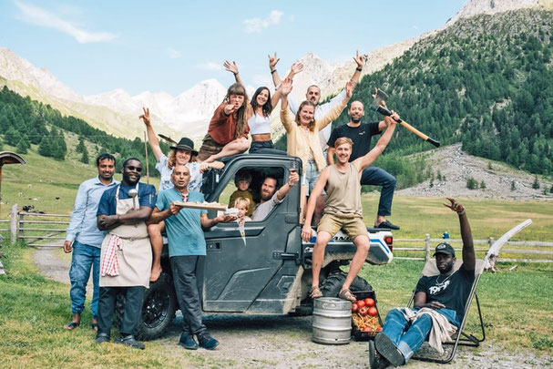 Fröhliches Eishof-Team posiert mit Werkzeug, Lebensmitteln und Getränken auf und um ein Fahrzeug, vor alpiner Bergkulisse im Pfossental in Südtirol.