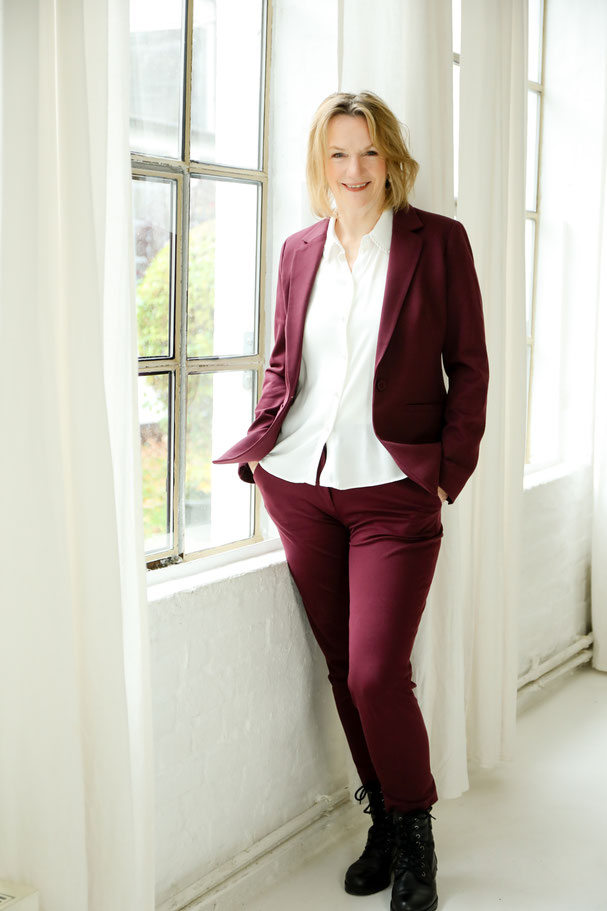 "Das Bild zeigt Sylke Damerau. Sie steht an einem Fenster und lächelt in die Kamera. Sie hat einen bordeaux farbenen Hosenanzug an."