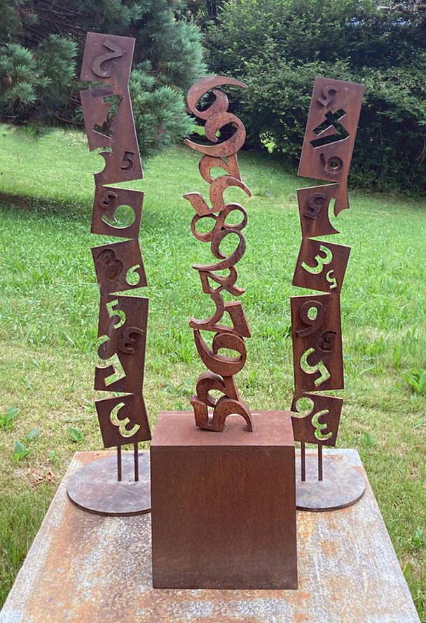 Two oxidized Corten steel sculptures from Paolo Grassi’s Steli 2.0 series, displayed outdoors among rocks and vegetation.