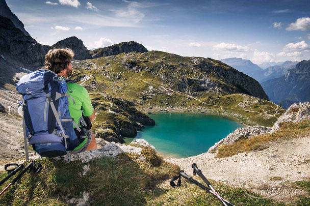 Wanderer sitzt auf einem Stein und blickt zu einem türkis farbenen Bergsee hinab