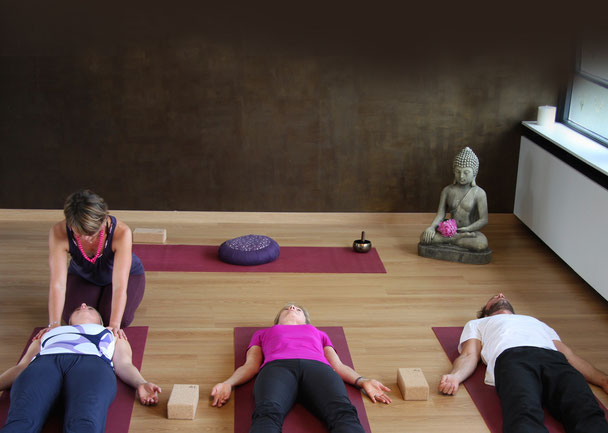 Somatic Yoga – Menschen in Savasana auf Matten; die Lehrerin berührt sanft die Schultern zur Erdung. Achtsame, anpassbare Praxis ohne anstrengende Stützhaltungen – fördert Entspannung, Körperbewusstsein und Nervensystem-Regulation.