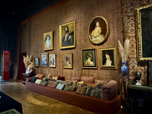 Eines der großen Sofas mit samtenen Kissen in warmen Farben, dahinter ein kleiner Teil der unzähligen Gemälde