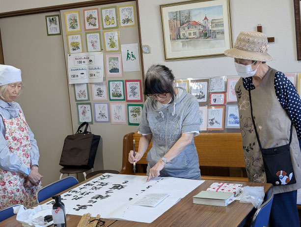 2026年5月31日（日）来週の礼拝案内を毛筆で準備しておられるのはりょうこさん。そして、それを見守るお二人は、近く、看板書き奉仕のデビューを果たすことに決まっている左から安佐子さんと光子さん！その日が来るのを楽しみに致しております。