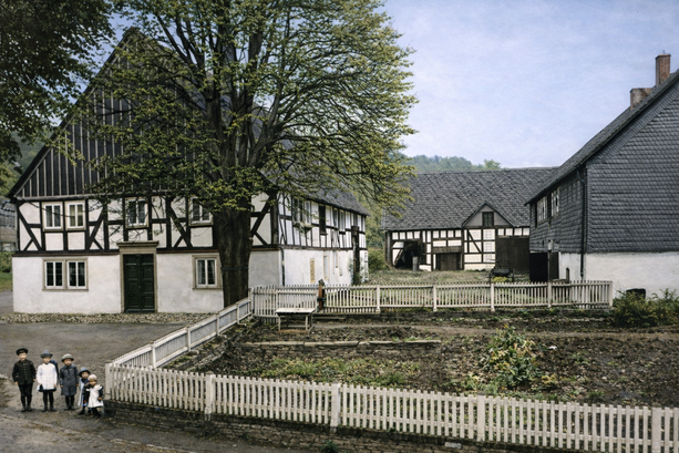 Foto um 1910 mit KI optimiert: Der Schulten-Hof in Eslohe mit Blick von der Pape-Straße aus auf den Innenhof, rechts das Wohnhaus mit Garten im Vordergrund, dahinter die Ökonomiegebäude (Stallungen, Remise und Scheune)
