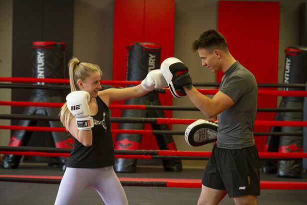 Junge Frau macht einen Fauststoß auf die Pratze ihres Trainers. Beide lachen und haben Spaß. Motivation, Fitness und Ausdauer. Selbstverteidigung und Persönliche Entwicklung. Gemeinschaft wird gefördert. 