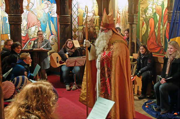 Nikolaus steht in der Kirche des Europaparks. Im Hintergrund sieht man die bunten Kirchenfenster. Um ihn herum sitzen Kinder und Jugendliche mit Musikinstrumenten und Liederzetteln in den Händen.