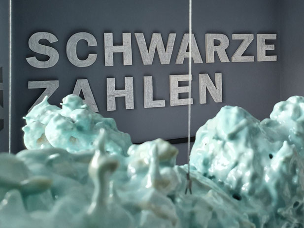 SCHWARZE ZAHLEN aus geweißtem Holz mit einem hölzernen schwarzen Unterstrich. Oliver Braig stellt uns immer wieder Fragen. Galeristin Andrea Oelmaier zeigt in der Ausstellung LOVE AND EROSION Arbeiten von Oliver Braig. Wer zahlt hier? Die Wände sind grau