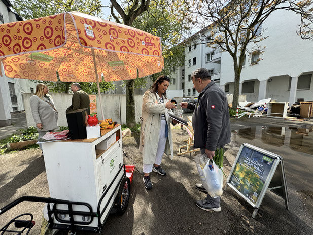 Ansprechpartnerin Feyza steht am Hallo Nachbar Infobike. Sie hält einem Nachbarn ein Glas mit Losen hin. Der Nachbar nimmt sich ein Los. 