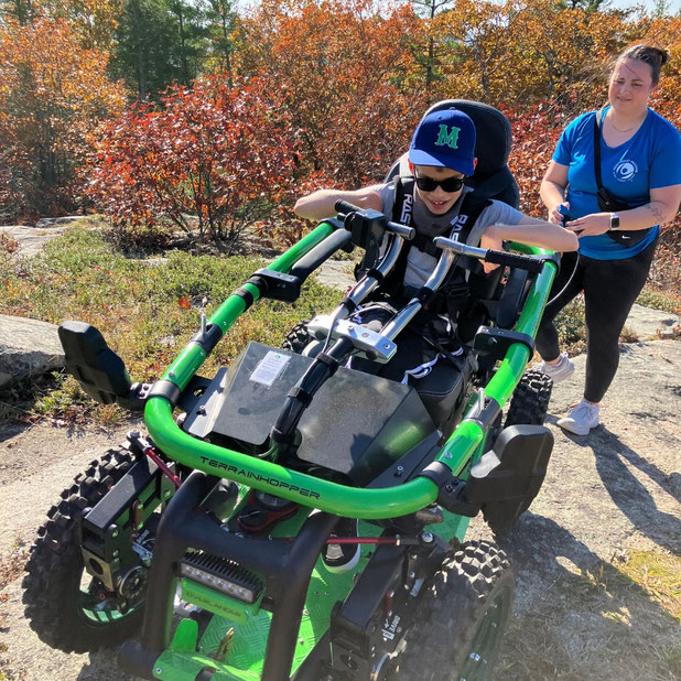 A young boy rides in a lime-green TerrainHopper, which is being driven with a tethered joystick by a Northeast Passage employee.