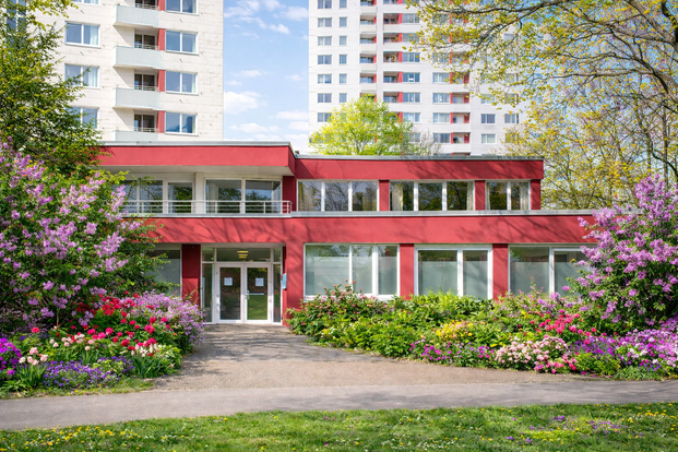 Rotes zweistöckiges Gebäude mit großen Fenstern und Eingangstür Praxis, umgeben von Bäumen und Grünflächen
