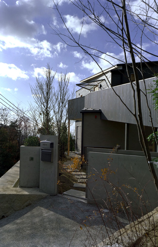 平賀敬一郎建築研究所が設計した、モダンなコンクリート門柱と木造建築が調和する住宅のアプローチ写真。踏み石が続く通路の先に見える建物の外観と、晴天の下の植栽。