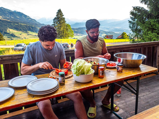 Zwei Teilnehmende des Integrationsprojekts Wanderglück schneiden auf der Terrasse der Selbstversorgerhütte im Allgäu gemeinsam Salat für das Abendessen.