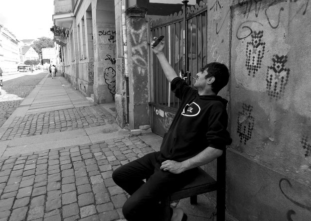 Fotografie von Elena Pagel, zeigt einen Mann der auf der Straße auf einem Stuhl Sitzt und ein selfie von sich macht. 