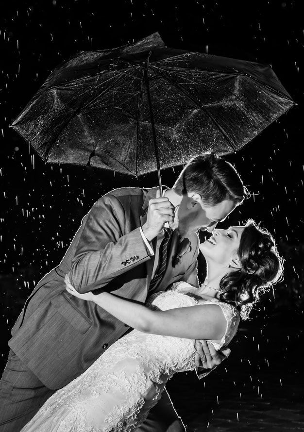 Black and white photo of bride and groom dancing in the rain. Groom is holding umbrella. Rain is backlit. 