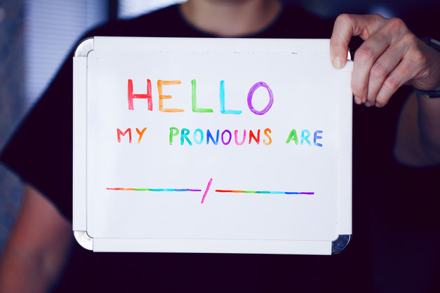 Person, die ein Tableau mit der Beschriftung "Hello, my pronouns are" hoch hält. Die Buchstaben haben die Farben der revidierten San Francisco Pride Flagge.