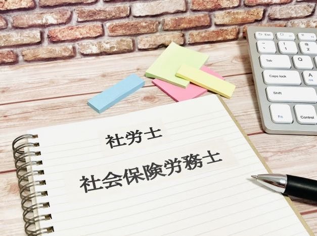 ノートに「社労士」「社会保険労務士」と書かれ、机の上にペン、付箋、キーボードが置かれている様子。