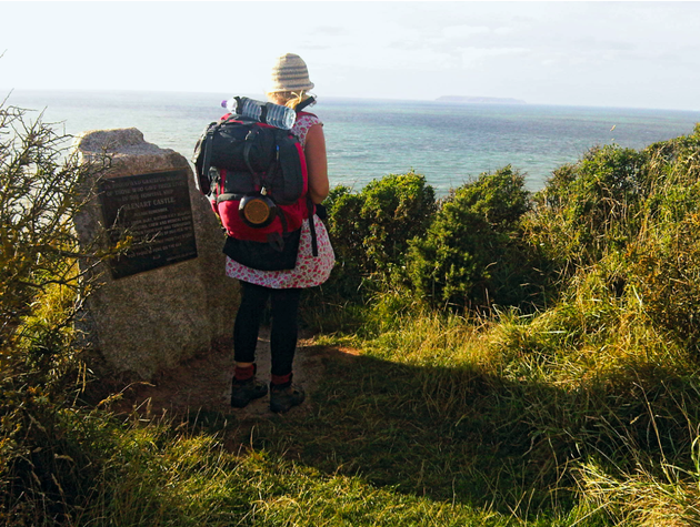 Frau mit Rucksack steht auf einem Hügel und guckt auf das Meer 