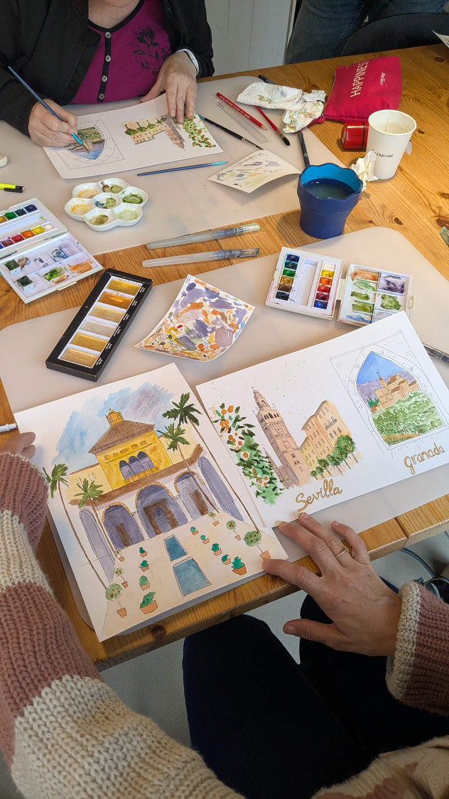 Aquarelle à grannulation Deux personnes peignent à une table avec des aquarelles, entourées de pinceaux et de palettes de couleurs. Les dessins représentent des paysages et des bâtiments. L'ambiance est créative et détendue