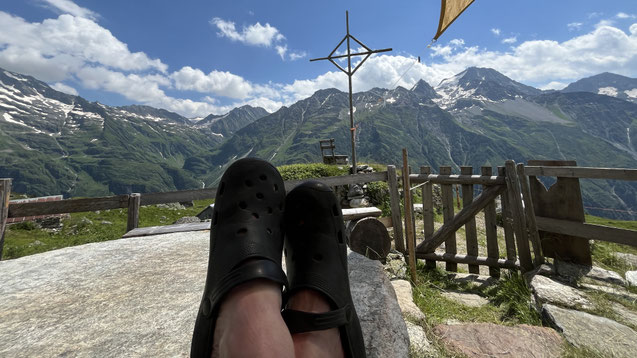 "Finde deine Ruhe: Dein Rückzugsort in den Schweizer Alpen." - Eine Person sitzt vor der Alphütte mit Blick über ein weites Bergpanorama