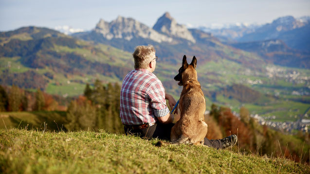 Ronald Kalbermatten und sein Hund Blue sitzen entspannt vor einer Alpwiese, Ronald schaut freundlich in die Ferne.