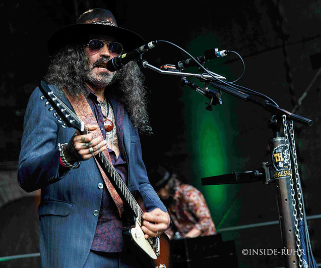 <img src="th-and-the-boneshakers-wattenscheid.jpg" alt="Musiker von TH & The Boneshakers spielt E-Gitarre auf der Bühne der Freilichtbühne Wattenscheid. Er trägt einen blauen Anzug, ein gemustertes Hemd und einen Hut.