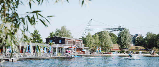 Wakeboard-Anlage am Allerpark Wolfsburg im Sommer – Blick durch Weidenäste auf die Wasserfläche, einen Wakeboarder und das historische Schloss im Hintergrund. Landschaftsfotografie Sebastian Bisch, Wolfsburg.