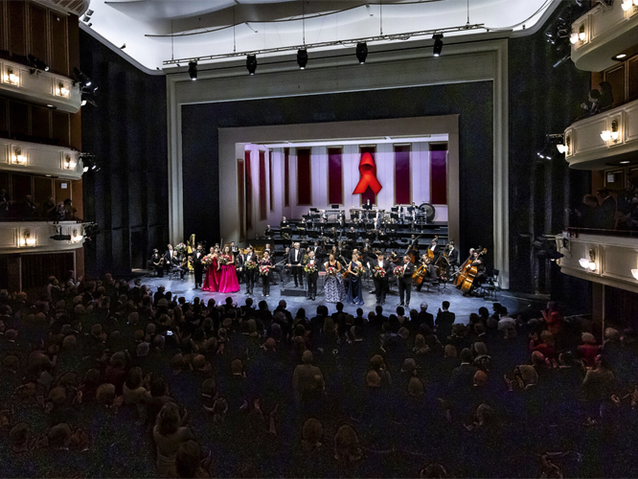 Bild: Mit Aidsschleife geschmückte Bühne im Opernhaus Düsseldorf mit Orchester und Sänger*innen 