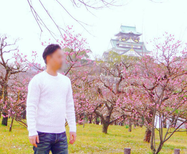 大阪梅田のメンズリラクゼーションサロン「forest hands」オーナー徹平のライフスタイル写真。満開のピンクの梅の花と大阪城天守閣を背景に佇む、白いニットを着たスマートな姿。