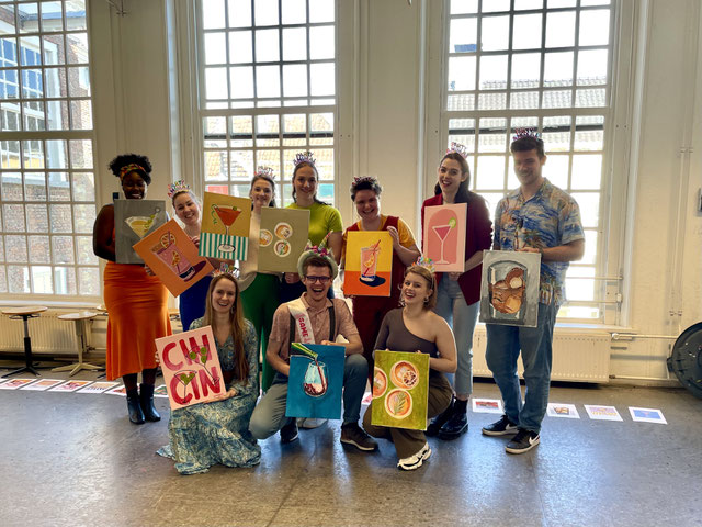 Vrolijke groepsfoto van een vrijgezellenfeest tijdens een schilderworkshop van Daphne Schrijver, met feesthoedjes, kleurrijke drankjes-schilderijen en een gezellige sfeer.