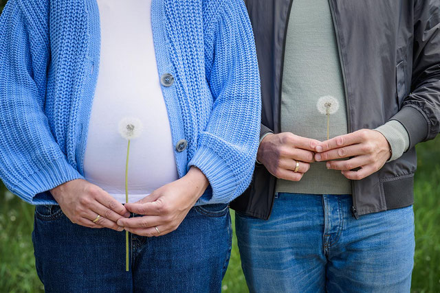 Schwangere Frau und Partner halten jeweils eine Pusteblume vor den Babybauch.