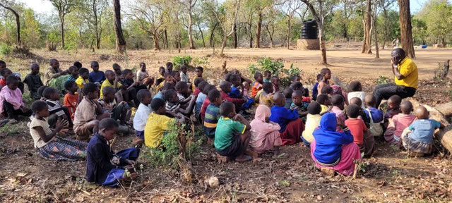 Eine große Gruppe bunt gekleideter Kinder die auf dem Erdboden in der Natur sitzen und einem Erwachsenen in gelbem Shirt zuhören