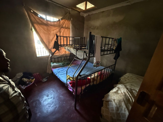 Schlicht eingerichtetes Schlafzimmer mit Metall-Hochbett und Matratzen; eine Person steht im Halbdunkel neben der Tür.