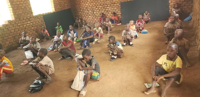 Eine Gruppe Kinder die in Reihen auf dem Lehmboden in einem Gebäude sitzen.