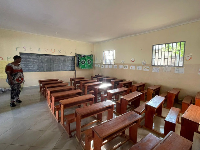 Ein leerer Klassenraum mit Holzbänken und einer Erwachsenen im Hintergrund