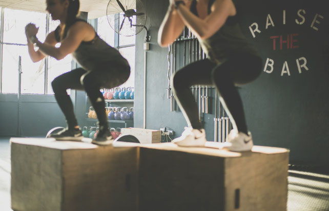 Zwei Frauen im Fitnessstudio machen Boxjumps. Beide sind in Schwarz gekleidet.