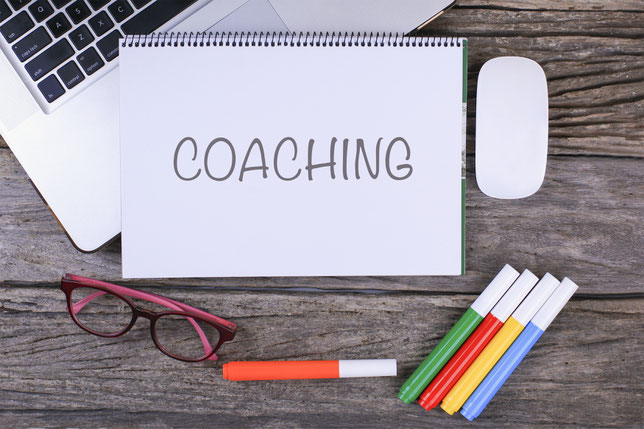 Holztisch mit Notizblock bezeichnet "Coaching", Laptop, Stifte und eine Brille