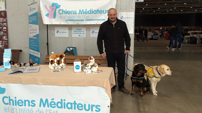 Jean-Pierre NEY assurant la promotion des CMUE lors d'un salon. A ses côtés : son chien de compagnie Nikita et un labrador en éducation au sein de l'association