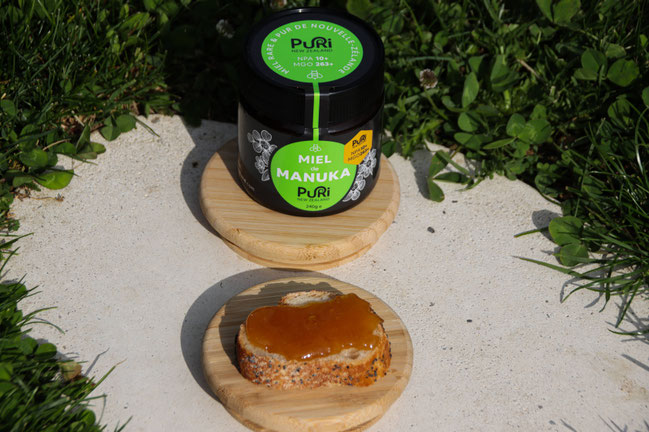 Miel de Manuka NPA 10+ Puri New Zealand en pot en verre de 240g au soleil sur un socle en bambou avec devant lui une tartine de pain grillé avec du Miel de Manuka 10+ et de l'herbe verte tout autour