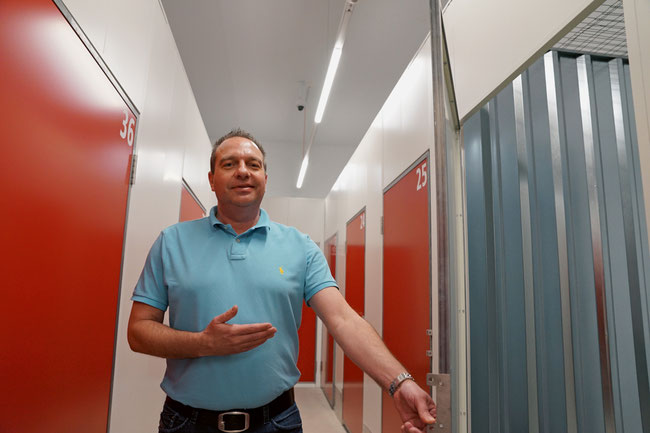 Marco Perlini vor einer geöffneten grösseren USB-Box., hinten an der Decke eine der Dauer-Videoüberwachungskameras.