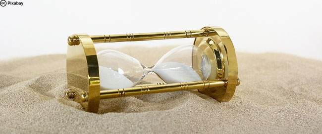 Eine goldglänzende Sanduhr liegt auf Sand. Bild von Annette auf Pixabay.
