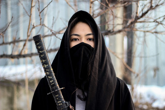 Eine asiatische Frau als Ninja gekleidet. Vor ihrem Körper hält sie ein Katana.
