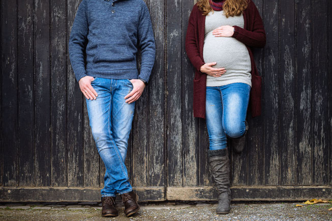 Schwangere steht neben ihrem Partner an einem Holztor und hält ihren Babybauch fest.