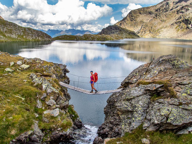 Hüttentrekking in den Alpen: Wanderin über eine Hängebrücke am Bergsee in hochalpiner Landschaft