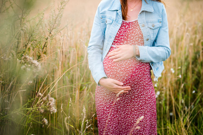Eine Schwangere steht im roten Kleid und Jeans-Jacke auf einer sommerlichen Wiese und hält dabei ihren Babybauch fest.