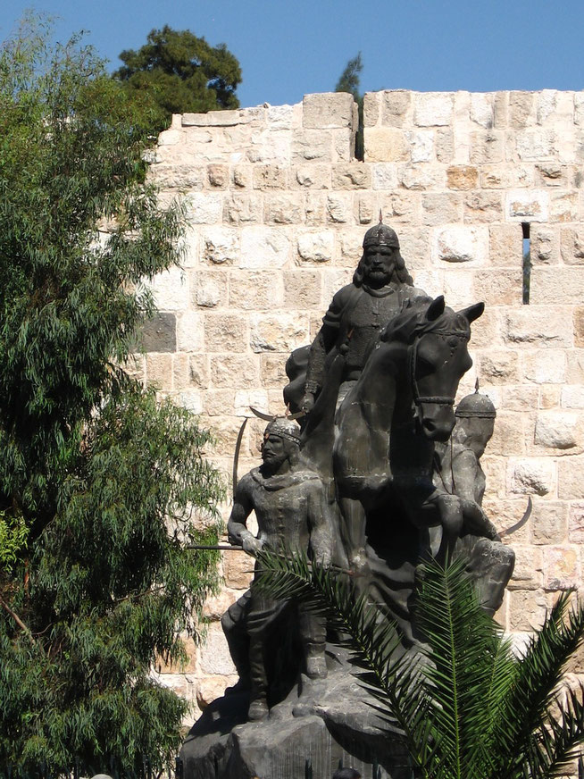 Bronze statue of a mounted warrior surrounded by armed figures, set against a stone wall with trees and clear blue sky.